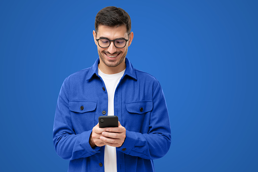 Young man looking at phone, standing isolated on blue background