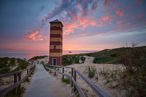 Lighthouse of Dishoek along the Dutch coast during a beautiful sunset. There is a path to the beach with a fence, the lighthouse is red and yellow striped. The sun is almost down and the sky has all the colors of the rainbow. A ship sails in the distance.