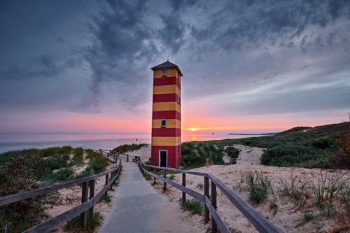 Lighthouse of Dishoek along the Dutch coast during a beautiful sunset. There is a path to the beach with a fence, the lighthouse is red and yellow striped. The sun is almost down and the sky has all the colors of the rainbow. A ship sails in the distance.
