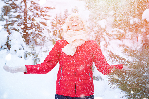 Happy woman in red jacket and warm knitted scarf catches snowflakes in winter forest on snowy day, sunlight