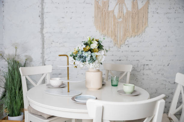 Stylish kitchen in white and brown wood. Style minimalism. Sink, table top, plants, pot, shelf for dishes. Bar stools, table. elegante cocina en blanco, colores pastel. estilo minimalista. jarrón con flores, mesa blanca, plantas, vasos, platos, platos. interior moderno con muebles blancos, mesa, paredes de ladrillo. loft apartamento .bright cocina en el estilo de la proven