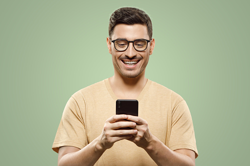 Portrait of young handsome man in beige t-shirt and eyewear, laughing at content he sees on smartphone screen, isolated on green background