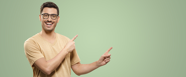 Banner of young smiling guy in beige t-shirt and eyewear, showing commercial offer on right, pointing to it with both hands, isolated on green background