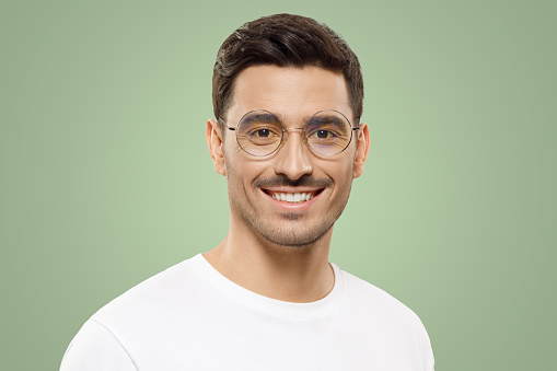Headshot portrait of young handsome guy wearing white t-shirt and round glasses, feeling confident and smiling happily, isolated on green background