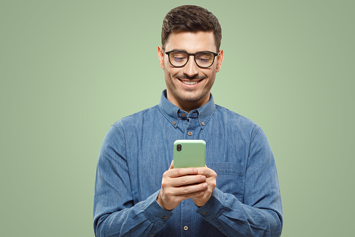 Young man wearing blue shirt and glasses, holding his smartphone and exchanging messages with frieends with smile, isolated on green background