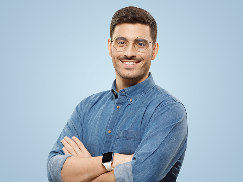 Young business guy in blue shirt and glasses, wearing black smart watch, standing with crossed arms and smiling happily, isolated on blue background