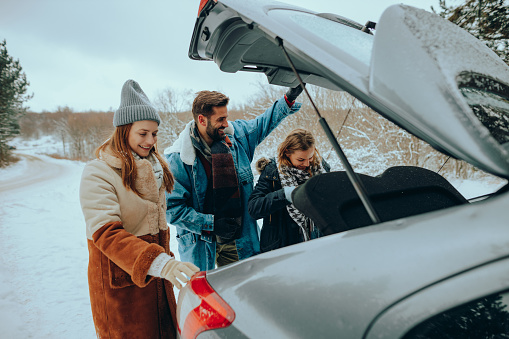 A happy group of friends, two young women and a one young man taking out winter gear from a car trunk on a snowy day in nature