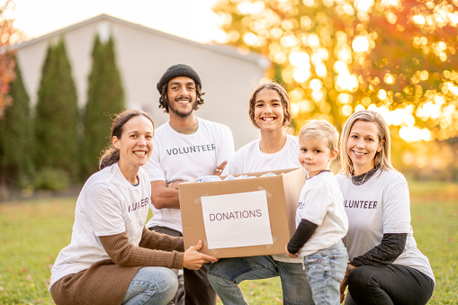 A small group of diverse volunteers huddle in closely together as they pose for a portrait during a community volunteer event.  They are each dressed casually in white t-shirts that read 
