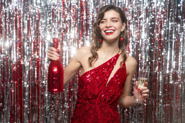 Dancing woman in red evening dress holding bottle and glass of champagne on tinsel background Dancing woman in luxury red sequins evening dress, with hairstyle and manicured nails, wearing red lipstick and christmas jewelry holding bottle and glass of champagne in hands on tinsel background woman in red dress stock pictures, royalty-free photos & images
