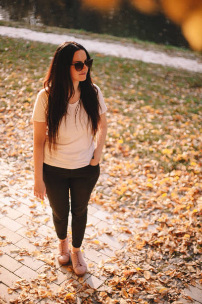 Full length portrait of happy stylish young woman in sunglasses looking away while standing in park during sunny weather in autumn stock photo