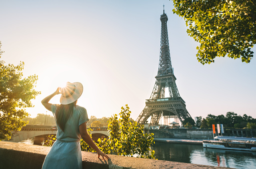Rear view of woman tourist in sun hat standing in front of Eiffel Tower in Paris at sunset. Travel in France, tourism concept. Holiday or vacation in Paris. High quality photo