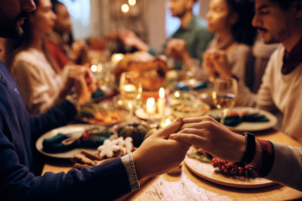 Close up of friends holding hands while praying during Thanksgiving meal. Close up of people saying grace before Thanksgiving dinner at dining table. saying grace stock pictures, royalty-free photos & images