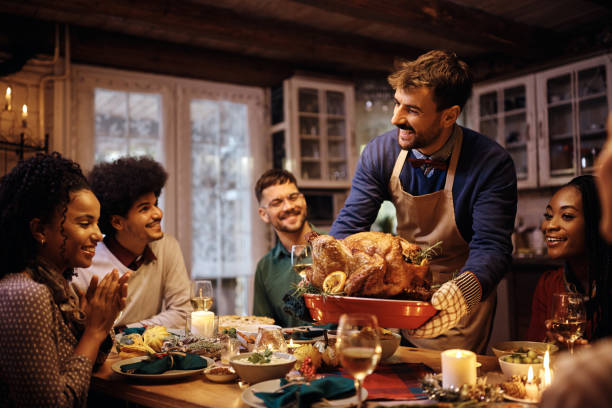 Young happy man serving Thanksgiving turkey to his friends at dining table. Happy man serving roasted turkey during Thanksgiving dinner with friends at dining table. social gathering stock pictures, royalty-free photos & images