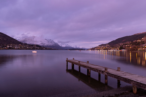 Queenstown, New Zealand, as viewed from the Francton beach. Dawn, after fresh snow has fallen on the mountains overnight. The sky starts to light up while the lights of the town are reflected in a peaceful Lake Wakatipu.