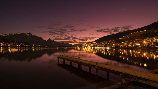 Queenstown, New Zealand, as viewed from the Francton beach.  The sillhouette of the mountains and the lights of the town are reflected in a peaceful Lake Wakatipu while the last light from a colorful sunset glows in the sky.