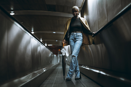 A low-key shot of a slender youthful black female with short hair painted white standing leaning on an umbrella in the middle of a travelator in a subway or a moving walkway of an underground crossing