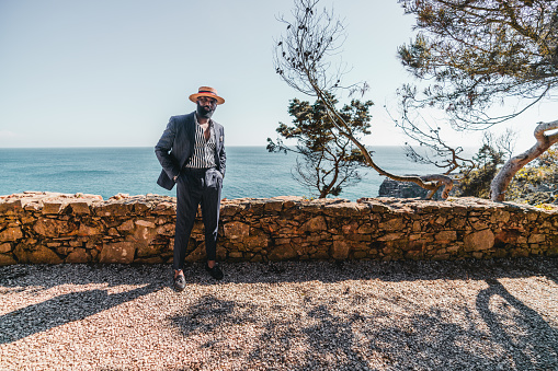 A wide-angle view of an elegant bearded mature black man in a straw hat and a tailored summer costume of a retro style with a striped shirt, standing on the shore next to the ocean and a stone fence