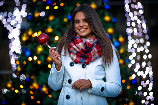 Portrait of a smiley woman in winter white jacket holding a caramel apple with bokeh lights behind her.