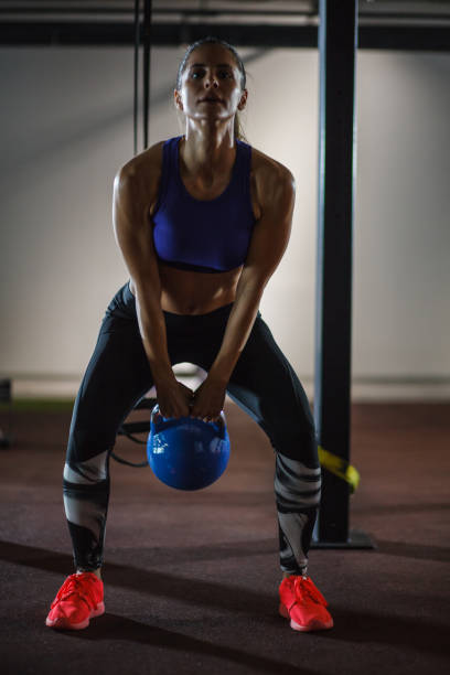 Muscular young female athlete concentrating while doing kettle bell swings to strengthen her upper and lower body muscles at the gym. She is looking at camera.