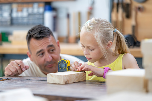 Father watching daughter measuring wooden plank with tape measure, medium shot. Family doing woodwork in carpentry workshop