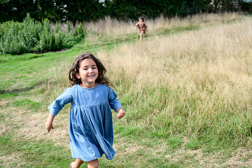 Focus on 6 year old girl in blue dress grinning at camera as she approaches and 5 year old sibling in background.