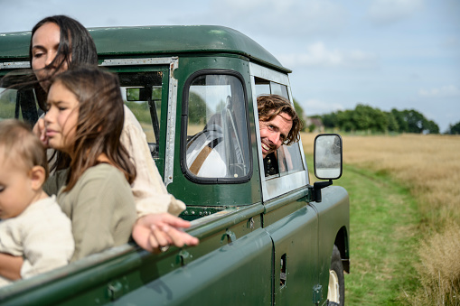 Candid portrait of market gardener grinning at camera from vehicle window while driving young families around as they learn benefits of local smallholding.