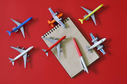 Notepad and fountain pen next to many toy models of passenger planes. The concept of choosing an air ticket, spotting, relocation or air travel. Red background. View from above. Close-up