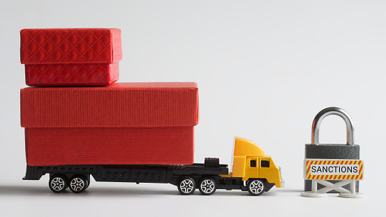 Toy truck with trailer loaded with boxes of goods stands next to prohibition sanction road sign and  padlock. White background. Ð¡oncept of trade sanctions and the impossibility of delivering. Close-up