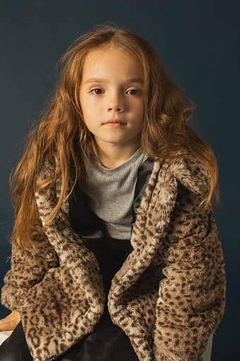 Redhead girl in warm winter clothes in studio on dark blue background. Little girl with long hair in fur coat. Portrait