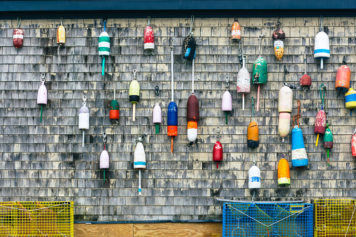 Traditional vintage lobster buoys hanging on the wall of a wooden shingled building with yellow and blue lobster pots on the ground in Portland Maine USA.