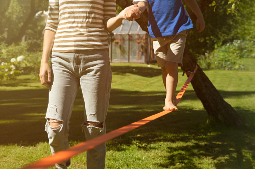 Mother holding kids hand walking on slackline training balance with bear foot on a warm sunny summer day in a green garden. Healthy living concept. Balance and help concept