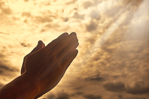 Hand praying with bright shining light on sky background. Religious and spiritual concept.
