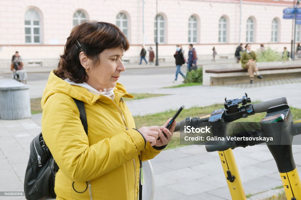 Mature woman in a jacket with a backpack rents an electronic scooter through a mobile application Mature woman in a jacket with a backpack rents an electronic scooter through a mobile application. City electric scooter rental service - kick sharing. Travel around the city on a scooter 60-64 Years Stock Photo