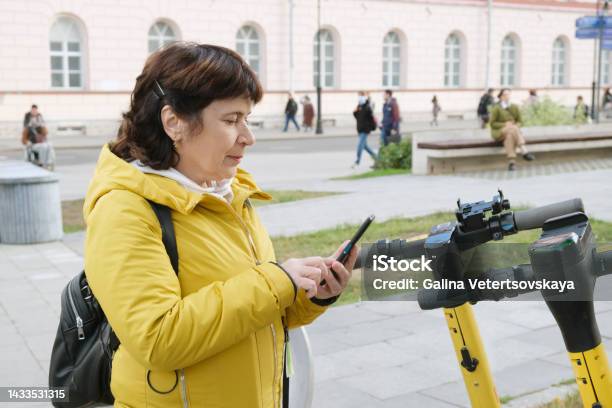 Mature Woman In A Jacket With A Backpack Rents An Electronic Scooter Through A Mobile Application Stock Photo - Download Image Now