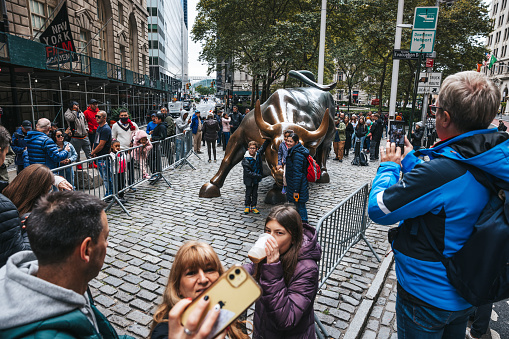 New York City, NY - October 5th 2022 : People wait in line to take a picture with the Charging bull sculpture in the financial district of downtown Manhattan in New York City USA. The bull is a three ton bronze sculpture located near Wall Street symbolizing New York's financial industry