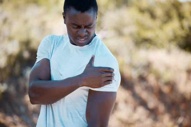 aktiver afroamerikanischer mann, der seine schulter vor schmerzen hält, während er während seines trainings in einer natürlichen umgebung an einer sportverletzung leidet. mann, der sich beim training im freien unwohl in seinem arm fühlt - shoulder pain physical injury muscular build stock-fotos und bilder