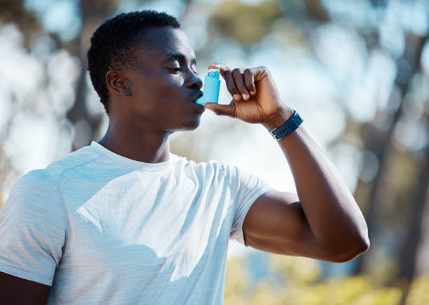Young african man taking a break from a workout to use his asthma pump. Fit athlete using his asthma inhaler during an asthma attack while exercising outside. Athletic man using a medical treatment One handsome young african american athlete using asthma pump after his daily workout routine outside. Dedicated black man taking care of his health and wellness. Endorsing a healthy lifestyle by exercising and taking his medication outside in a forest asthmatic stock pictures, royalty-free photos & images