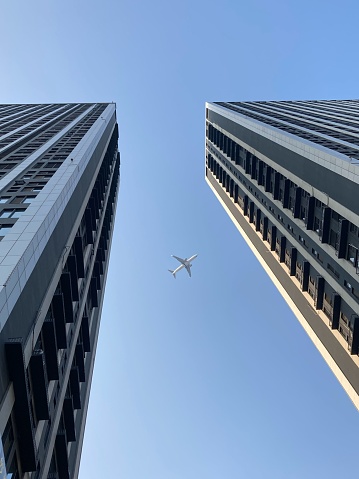 A vertical undershot of two skyscrapers with the airplane flying over the blue sky background