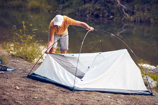 Man camping in nature, setting up the tent for overnight staying near forest river.