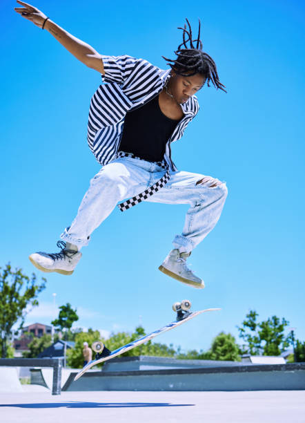 jeune garçon cool qui exécute des figures sur son skateboard au skatepark. jeune homme afro-américain concentré sautant pour retourner son skateboard en l’air. jeune patineur élégant profitant du parc - hip hop hipster afro men photos et images de collection