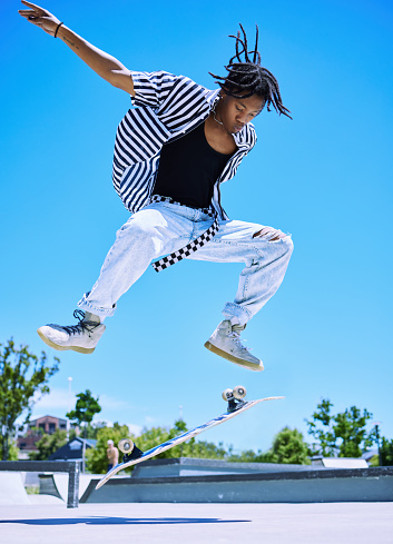 Cool young boy performing tricks on his skateboard at the skatepark. Focused young African American man jumping to flip his skateboard in the air. Stylish young skater enjoying the park