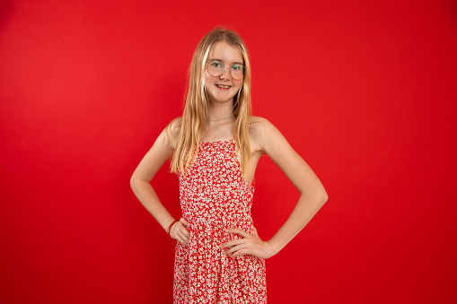 Portrait of smiling beauteous teenage girl with long fair hair wearing light summer dress, glasses, holding hands on waist, looking at camera, posing on red background. Studio. Adolescence, childhood.