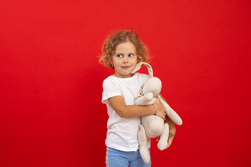 Portrait of cheerful blue-eyed little girl with short curly fair hair wearing white T-shirt, jeans holding white soft toy rabbit, looking aside, standing on red background. Studio. Present, childhood.