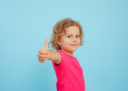 Portrait of smiling blue-eyed little girl with curly fair hair wearing pink T-shirt, looking, showing thumb up sign on blue background. Studio. Advertisement, approval, emotions, feelings, copy space.