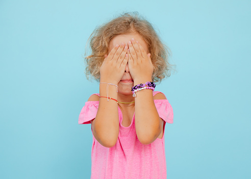 Portrait of upset scared little girl with short curly fair hair, wearing pink jumpsuit, bracelets, covering eyes with hands hiding face on blue background. Emotions, fears, hide-and- seek, studio.