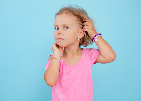 Portrait of cherubic blue-eyed little girl titivating touching short curly fair hair, wearing pink jumpsuit, bracelets, looking at camera, posing on blue background. Childhood, copy space, studio.