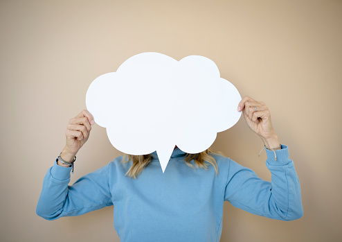 beautiful young woman with curly blond hair stands in front of brown wall and holds white speech bubble, thought bubble