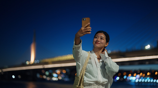 A young female tourist is taking selfies with her smart phone in the city at night.