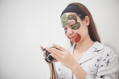 Asian woman with cosmetic mask on her face, looking at photos in digital camera, Wellness at home.