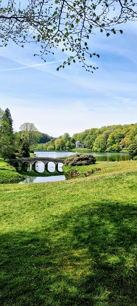 A vertical shot of a park with an old stone bridge over a river with beautiful trees in the background